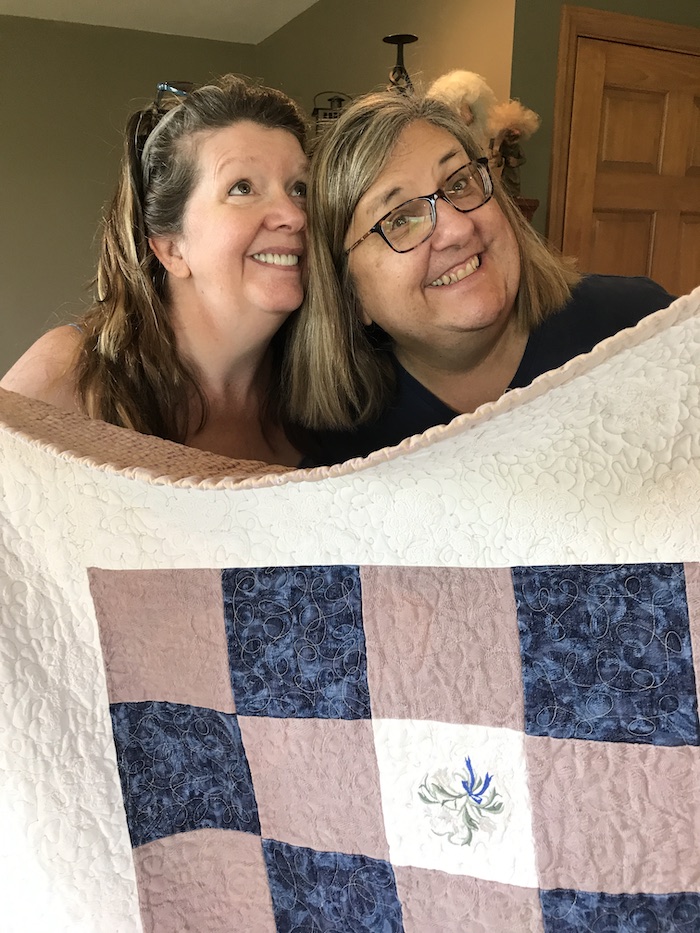 Paola Jo and Quilter smiling for camera as they hold up a blue and pink quilt